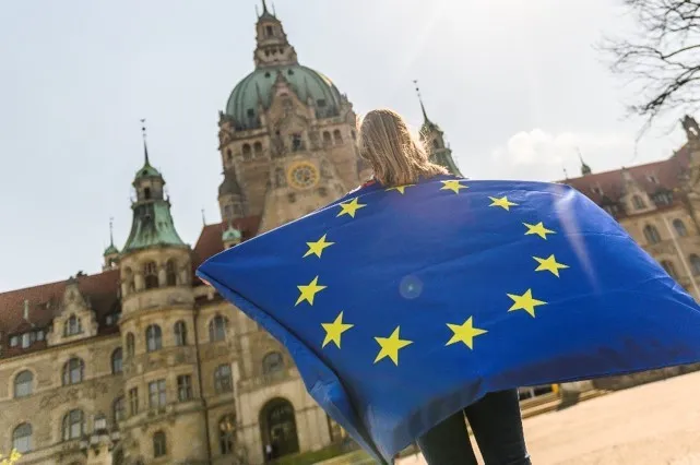 Ein Mensch blickt auf das Neue Rathaus von Hannover, das im Hintergrund des Bildes zu sehen ist. Der Mensch steht mit dem R&uuml;cken zur Kamera und hat eine Europaflagge &uuml;ber den R&uuml;cken gespannt. Der Fokus des Bildes liegt auf der Flagge.